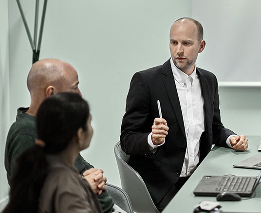 Man with pen raised making a point while sat at a meeting table with two colleagues